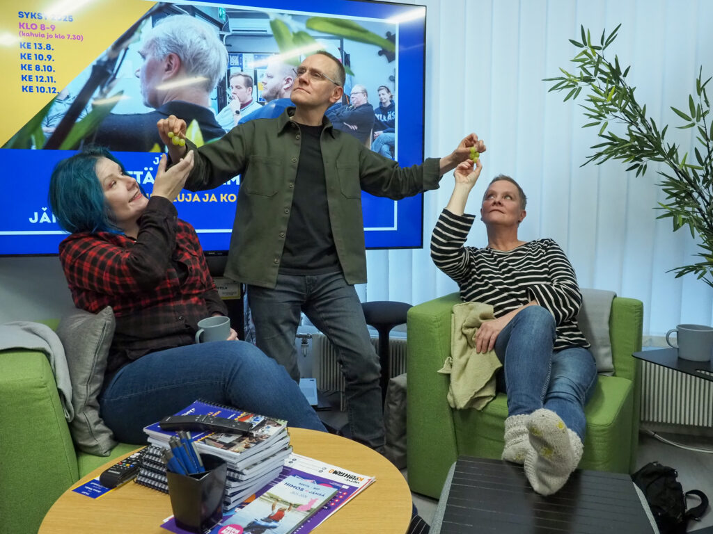 Kolme ihmistä, kaksi naista istumassa ja keskellä mies antamassa naisille viinirypäleitä. Paikkana Jämsä Tehdas -etätyötilan sohvaryhmä.
