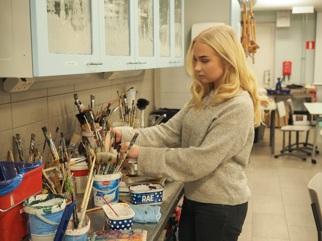 Nuori nainen Jämsän lukiolla kuvataideluokassa valitsemassa siveltimiä pöydältä.