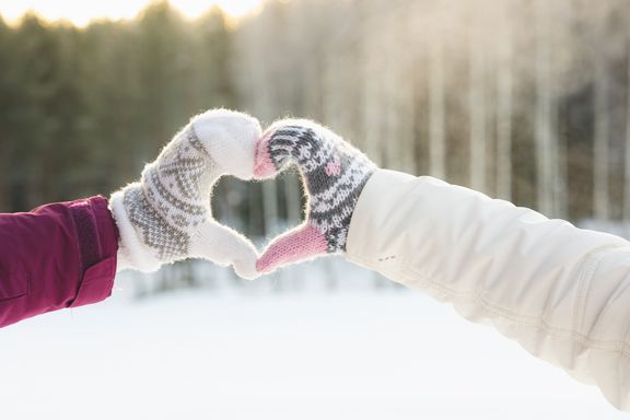 Kahden ihmisen kädet muodostavat sydämen talvimaisemassa, lapaset käsissä