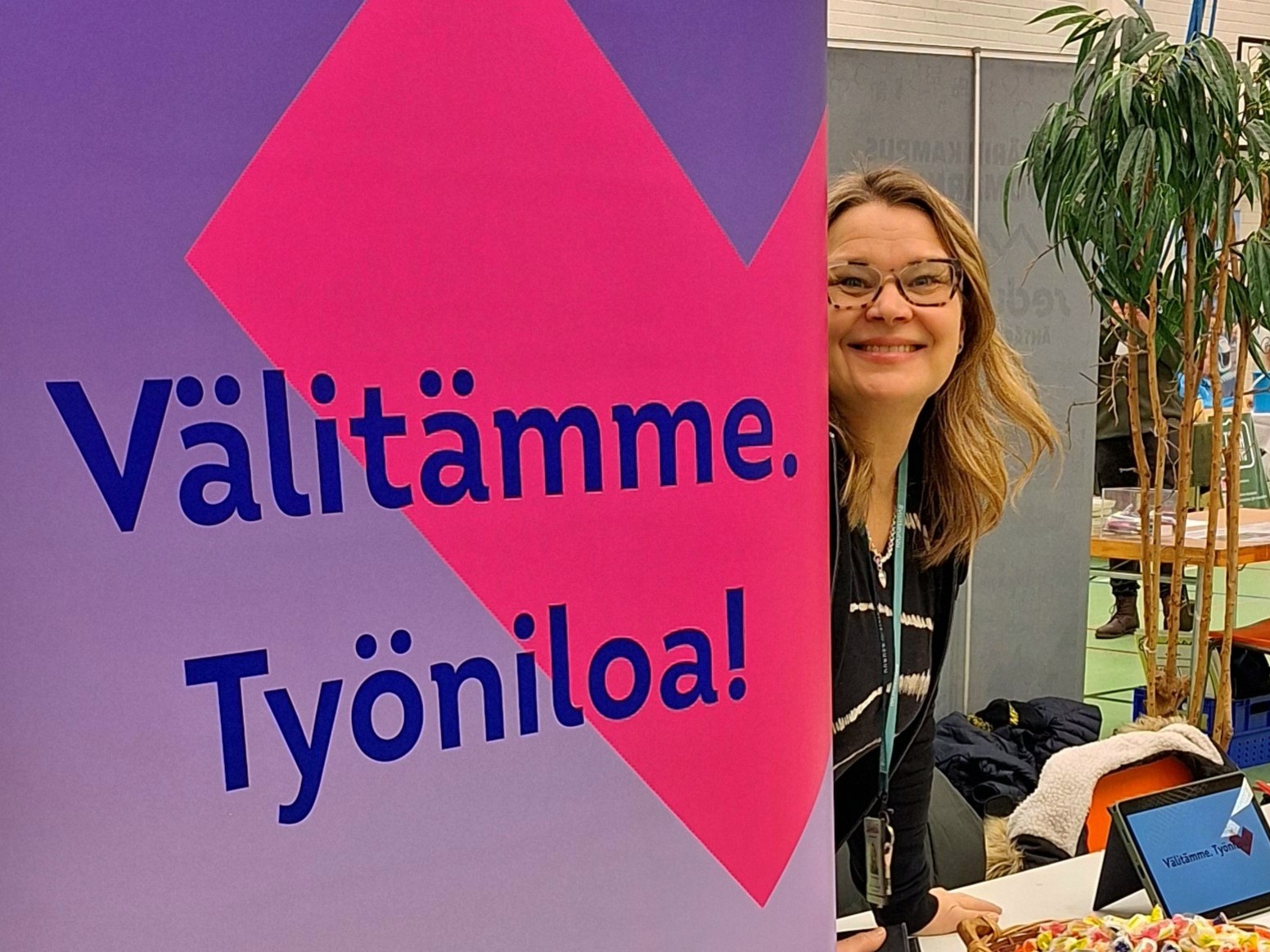 Henkilö kurkistaa messujulisteen takaa. Julisteessa lukee Välitämme työniloa.