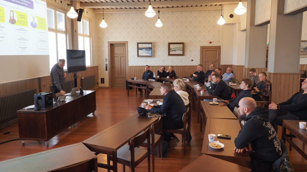 Ihmisiä istumassa pöytien äärellä salissa, Jokivarren puukoululla.