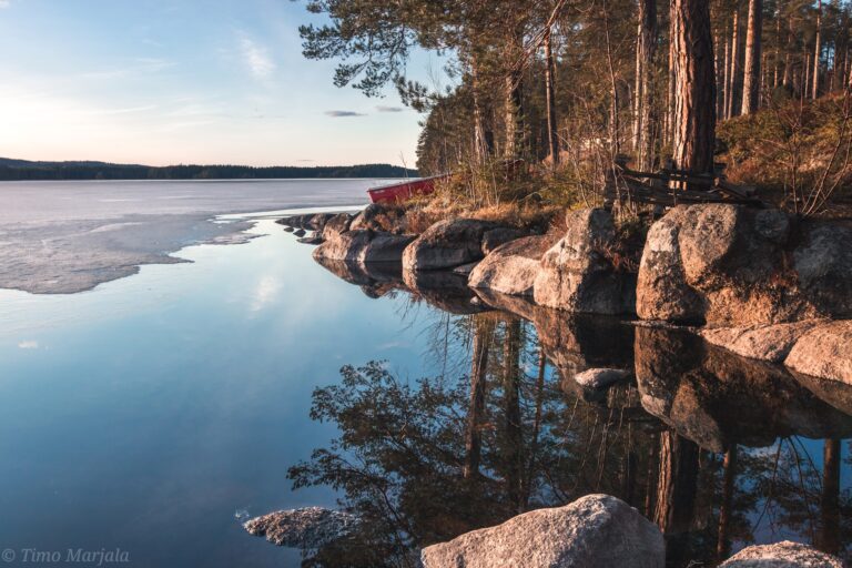 Keväinen maisema, kivinen ranta, puita ja järvi joka osittain jäässä.
