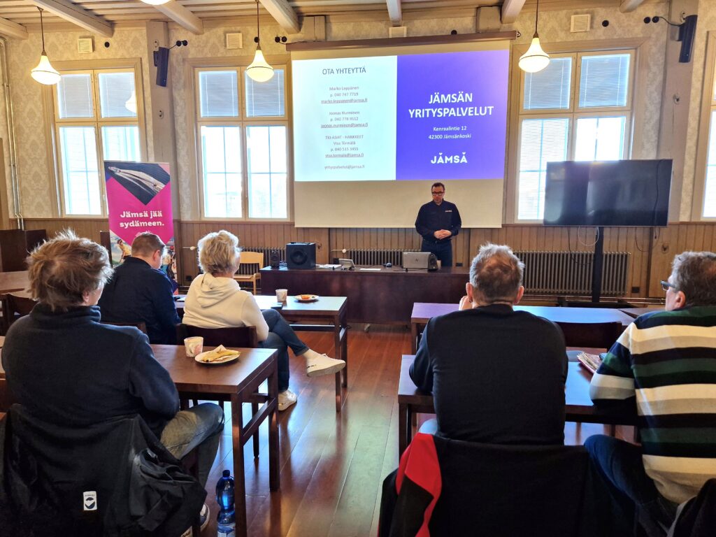 Ihmisiä istumassa pöytien äärellä salissa, paikkana Jokivarren puukoulu.