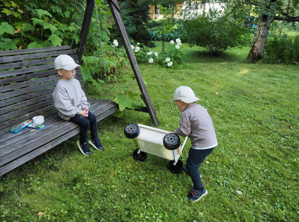 Kaksi lasta ulkona, toinen istuu keinussa ja toinen työntää lasten kottikärryjä.
